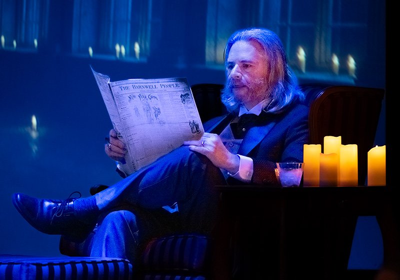 Male actor sitting on stage reading a newspaper lit by candles.